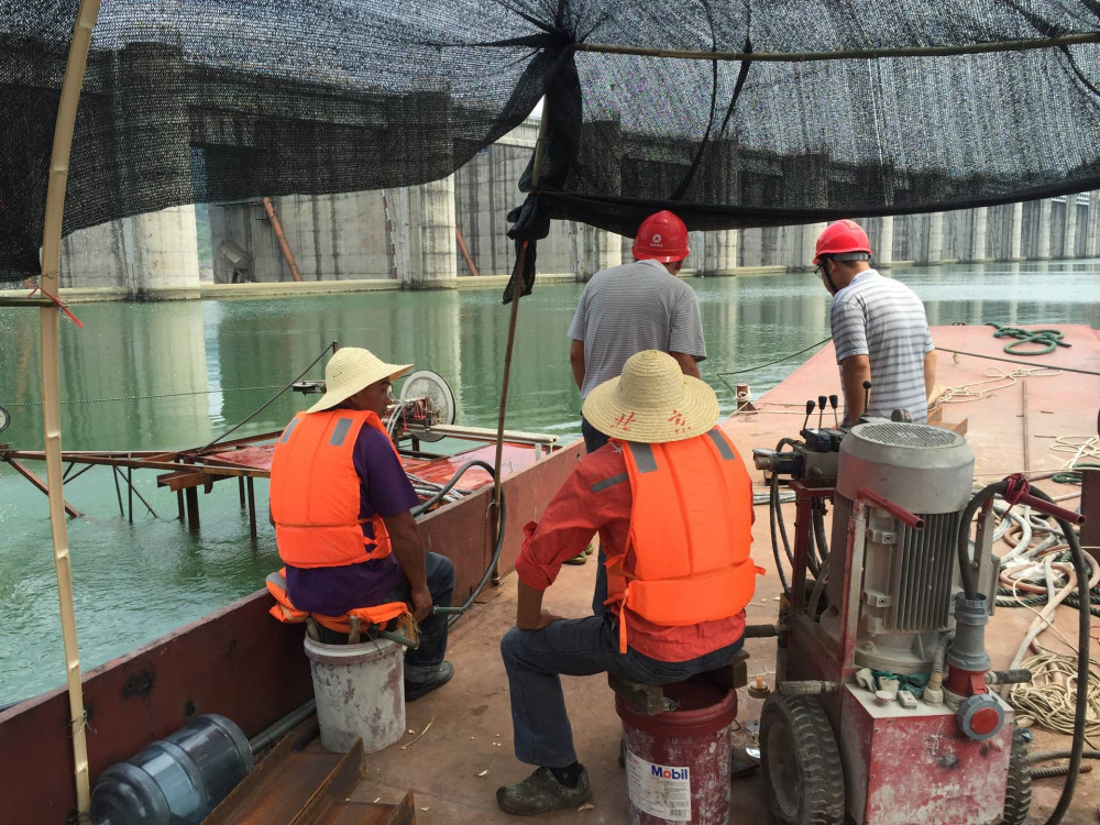 水下切割作业平台实拍：驳船甲板上穿救生衣、戴安全帽的施工人员操作切割设备，液压泵站与导向滑轮在甲板就位，远处为水库大坝